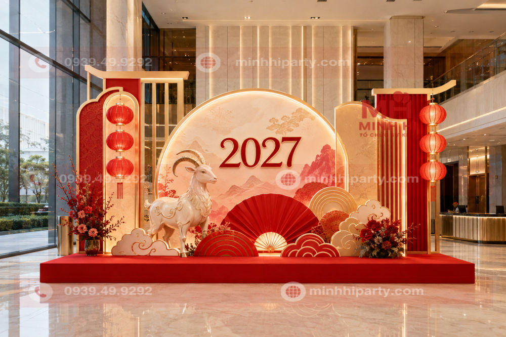 Mẫu trang trí Tết 2027 cho chung cư cao cấp với backdrop đỏ, vàng champagne, đèn LED và linh vật dê tại sảnh.
